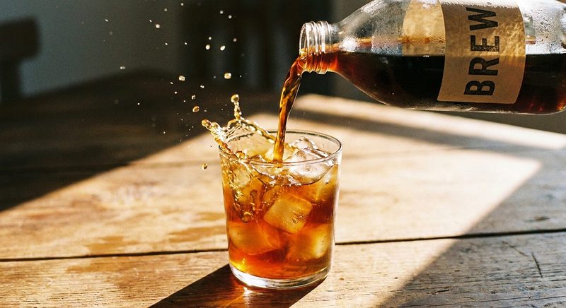 Cold brew pouring from a bottle into a glass of ice on a sunlit wooden table