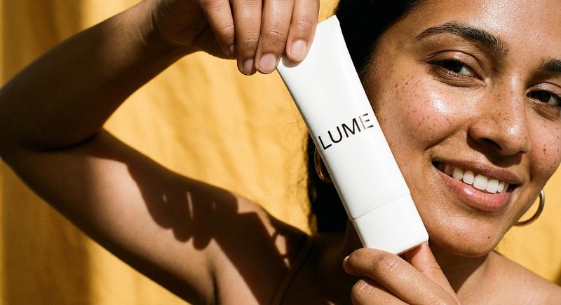 Smiling woman in golden light holding a white skincare tube beside her face