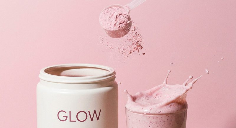 Scoop of pink protein powder pouring into a shaker glass beside a white tub