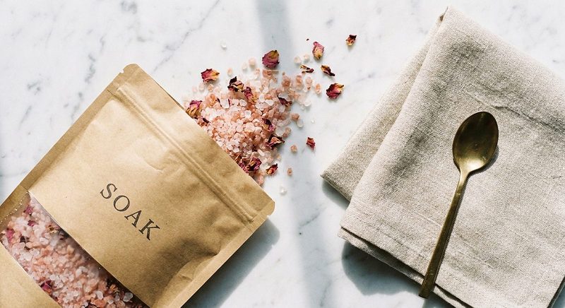 Kraft pouch spilling pink bath salts and rose petals on marble beside a spoon